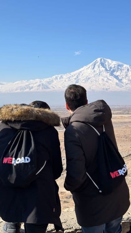 Due persone di un viaggio di gruppo WeRoad, viste da dietro, guardano una montagna innevata in lontananza sotto un cielo sereno.