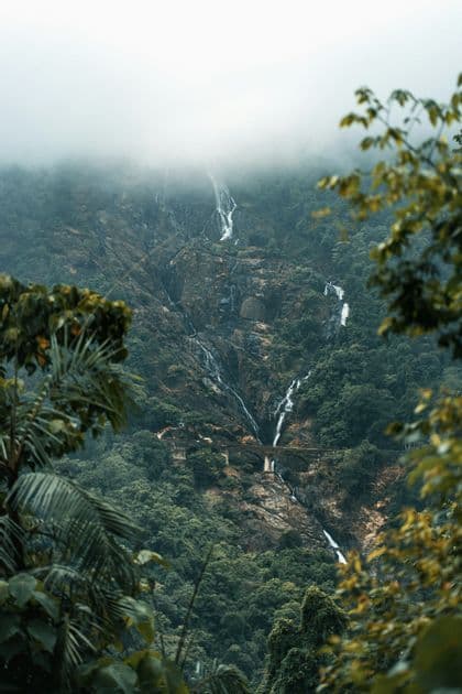 Le cascate scendono lungo un fianco di montagna roccioso e boscoso, avvolto nella nebbia, con un ponte ad arco visibile in lontananza.