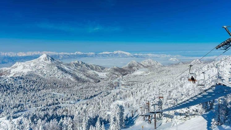 Una vista panoramica da una seggiovia mostra una vasta catena di montagne e foreste innevate sotto un cielo azzurro brillante.