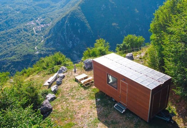 Una tiny house in legno con pannelli solari, situata su un fianco di montagna, con vista su una lussureggiante valle verde.