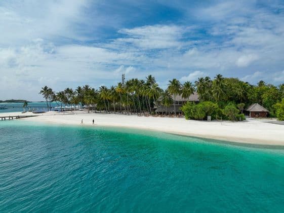 Una vista aérea de una isla tropical con playa de arena blanca, agua turquesa y palmeras bajo un cielo parcialmente nublado.