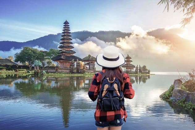 Una mujer con sombrero y mochila observa un complejo de templos de varios niveles a orillas de un lago, con montañas al fondo.