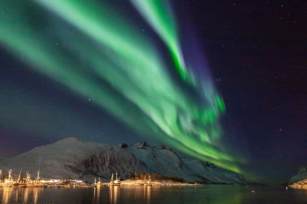 The green aurora borealis glows in the starry night sky above snow-covered mountains and a harbor with boats.