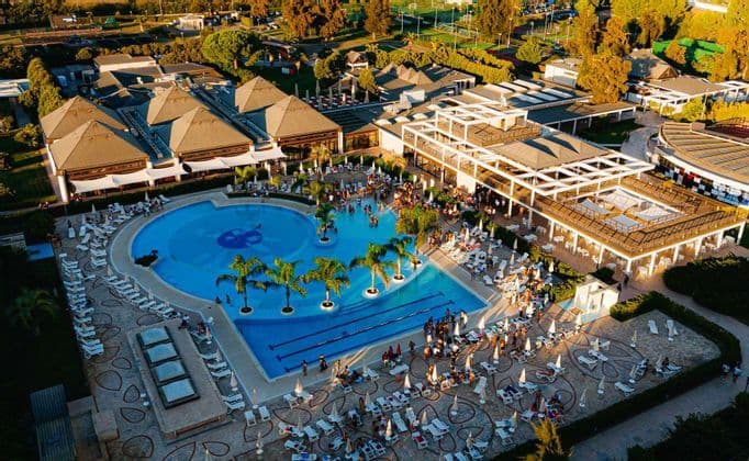 Vista aerea di un grande resort con persone che si godono una piscina circondata da palme e sedie a sdraio al tramonto.