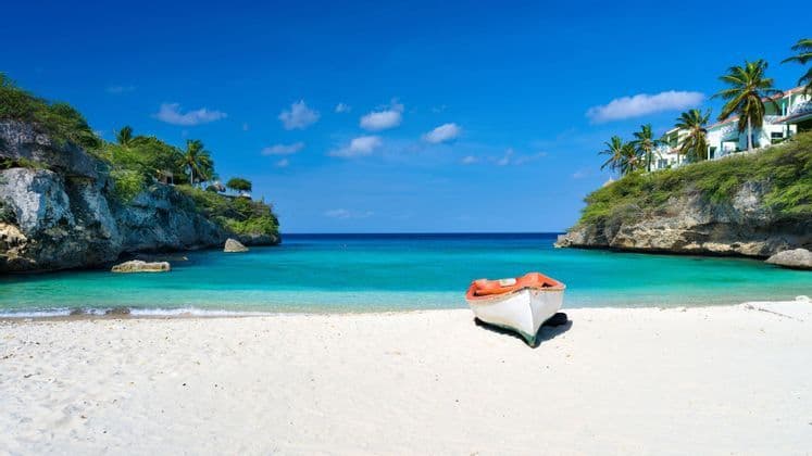 Una piccola barca si trova su una spiaggia di sabbia bianca in una caletta con acqua turchese e scogliere sovrastate da palme.