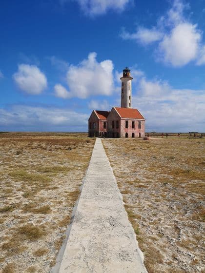 Un sentiero in cemento conduce a un vecchio faro in un campo arido sotto un cielo azzurro nuvoloso, con un viaggio di gruppo WeRoad nelle vicinanze.