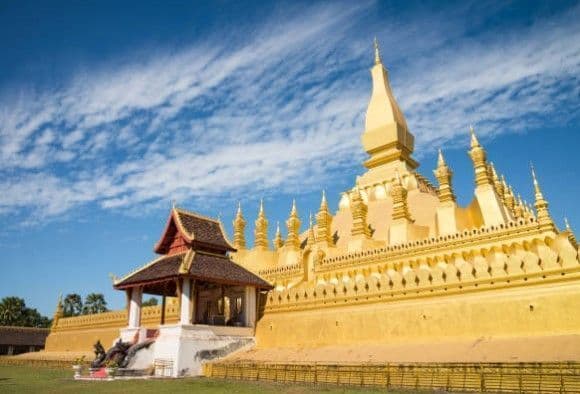 Un grand stupa doré aux flèches complexes se dresse à côté d'un petit pavillon sur une pelouse verte sous un ciel bleu et nuageux.
