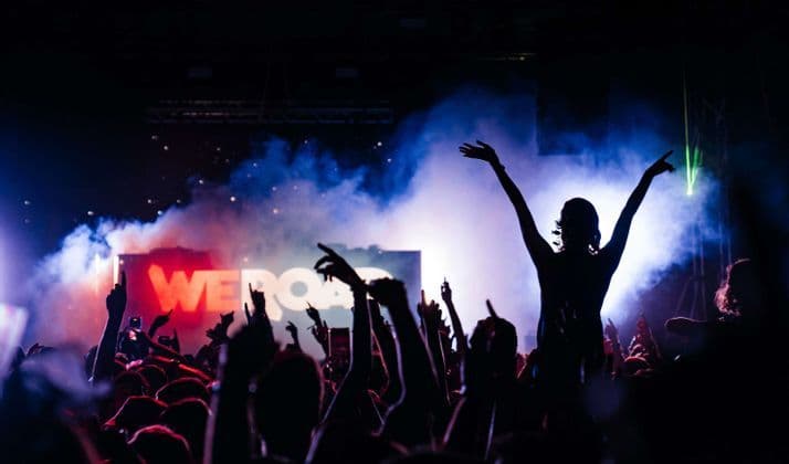 Una folla di persone in silhouette con le mani alzate a un concerto WeRoad con luci di scena viola e fumo.