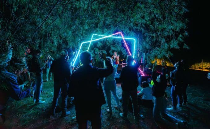 Un viaggio di gruppo WeRoad assiste a uno spettacolo musicale all'aperto di notte sotto gli alberi, davanti a un palco con luci al neon geometriche.