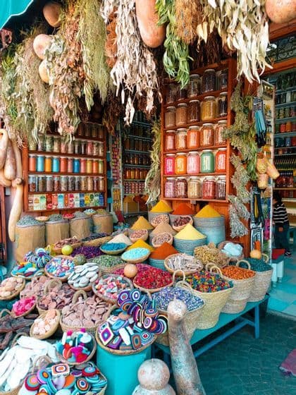 Ein Marktstand präsentiert farbenfrohe Gewürze in Körben und pyramidenförmig aufgeschichtet, dazu Gläser in Regalen und getrocknete Kräuter, die von oben herabhängen.