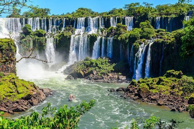 Una barca turistica naviga su un fiume ai piedi di un'enorme cascata a più livelli, circondata da rigogliose scogliere verdi.