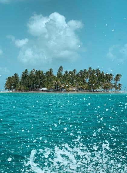 Un'isola tropicale coperta di palme vista dal mare turchese, con l'acqua che schizza in primo piano.