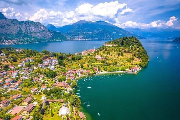 Veduta aerea di una città su una penisola verde che si protende in un lago blu, con montagne sullo sfondo.