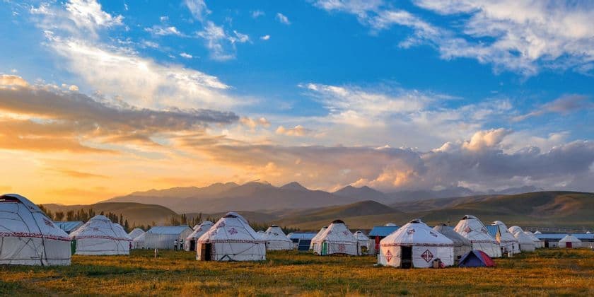 Un campamento de yurtas blancas se asienta en un campo de hierba con montañas al fondo bajo un cielo al atardecer.