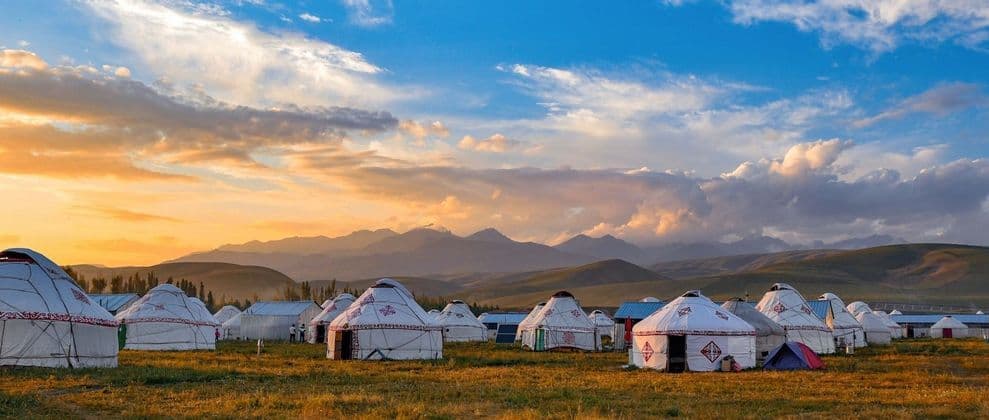 Un accampamento di yurte bianche in un campo erboso con una catena montuosa visibile sullo sfondo sotto un cielo al tramonto.