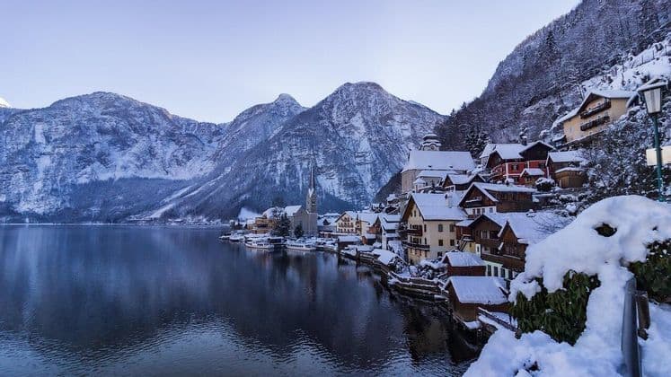 Ein schneebedecktes Dorf mit einer Kirche, eingebettet an einem ruhigen Seeufer am Fuße großer Berge unter einem klaren Himmel.