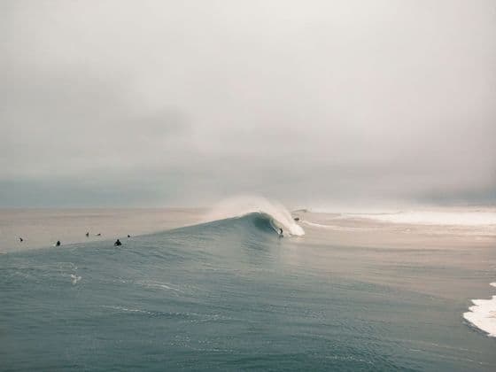 Un surfista cavalca una grande onda tubante nell'oceano, mentre altri surfisti attendono sotto un cielo nuvoloso.