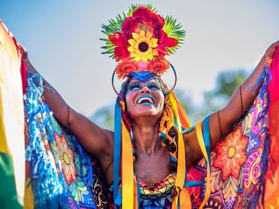 A smiling woman in a vibrant carnival costume with a large floral headdress celebrates with her arms raised.