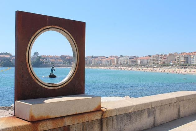 Una escultura de metal oxidado con una abertura circular enmarca la vista de una estatua en el mar frente a una playa concurrida.