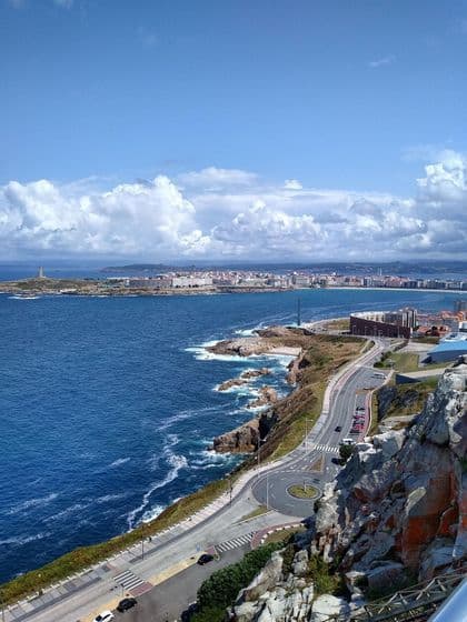 Vista elevada de una carretera costera serpenteante junto al océano azul, con una ciudad y una torre lejana bajo un cielo nublado.