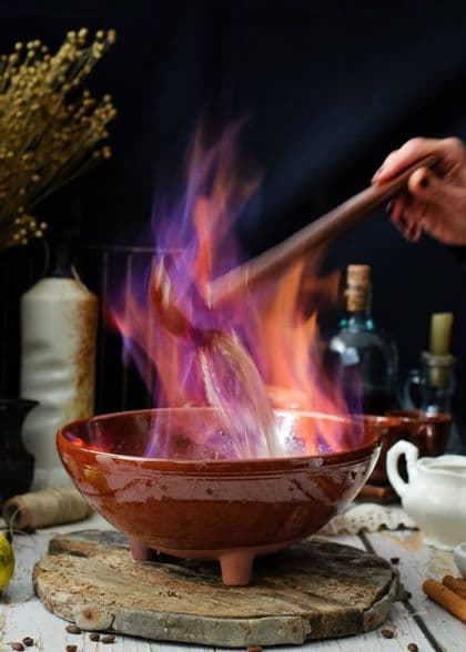 Una mano remueve un líquido flameante púrpura y naranja en un cuenco de terracota con un cucharón de madera.
