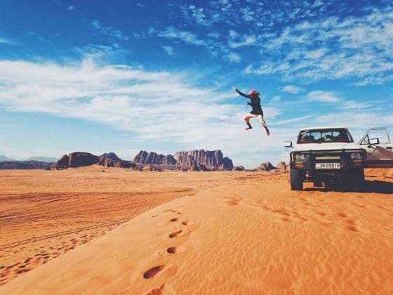 A person jumps in the air on an orange sand dune in the desert, next to a white pickup truck, with mountains in the background.