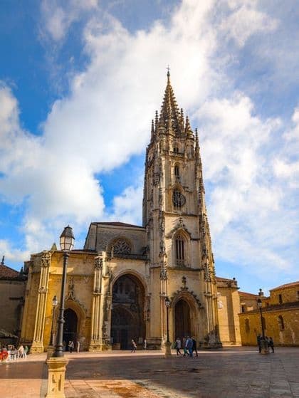 Una ornamentada catedral gótica con una alta aguja puntiaguda, vista desde una plaza pública bajo un cielo azul y nublado.