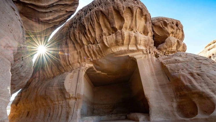 Un'antica tomba scavata in una scogliera di arenaria, vista dal basso, con un'esplosione di luce solare che filtra tra le rocce.