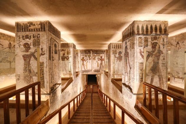 In einem altägyptischen Grab führt ein hölzerner Steg mit Geländer zwischen Säulen, die mit farbenprächtigen Hieroglyphen bedeckt sind, hinab.
