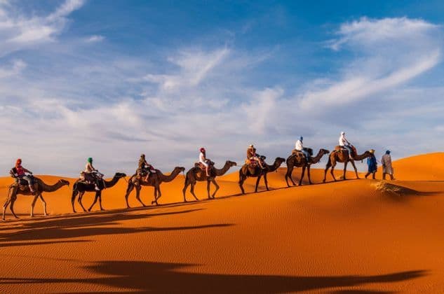 A WeRoad group trip rides a line of camels over orange desert sand dunes under a partly cloudy sky.