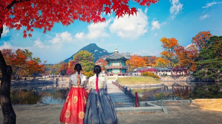 Due donne in abiti tradizionali hanbok guardano un lago verso una pagoda circondata da un vibrante fogliame autunnale.