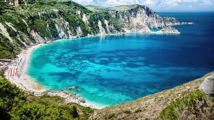 Una vista dall'alto di una cala appartata con acqua turchese, una spiaggia sabbiosa con persone e ripide scogliere verdi e bianche.