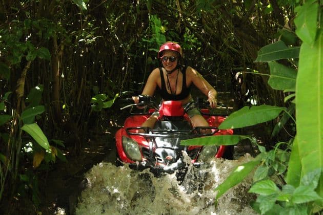 Eine Frau mit rotem Helm fährt ein rotes Quad und spritzt durch Wasser in einem dichten, sonnendurchfluteten Dschungel.