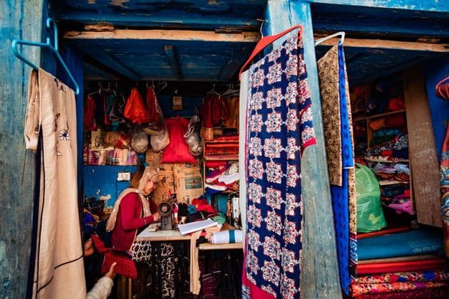 A woman works on a sewing machine inside a small, blue-painted stall filled with colorful fabrics and hanging garments.