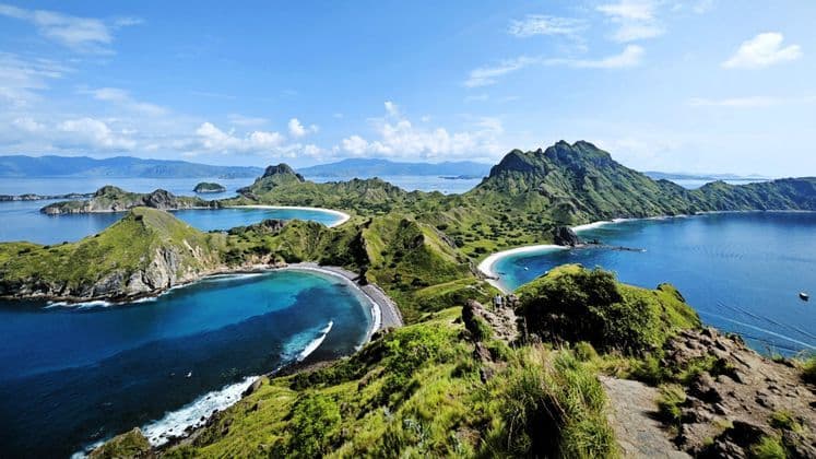 Eine Vogelperspektive auf schroffe grüne Inseln mit Sandbuchten, wo zwei Personen auf einem Grat den türkisfarbenen Ozean überblicken.