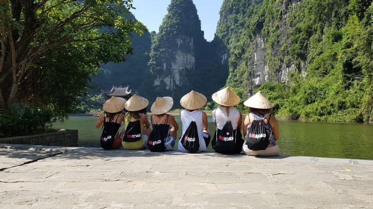 Un groupe WeRoad est assis sur une corniche rocheuse, coiffé de chapeaux coniques et admirant une rivière entourée de montagnes verdoyantes.