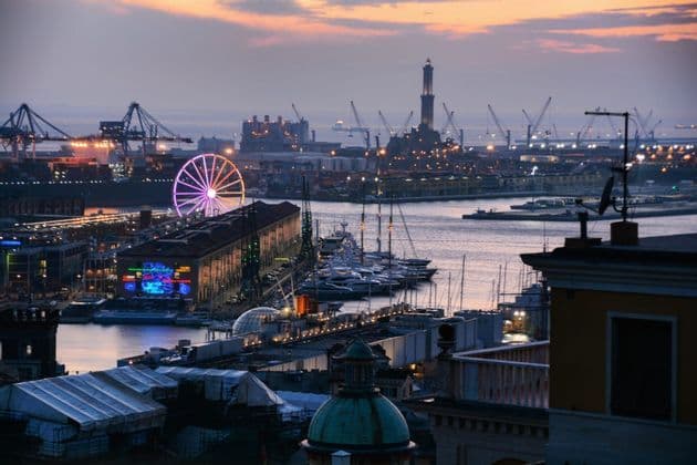 Una vista aerea di un porto al crepuscolo, che mostra una ruota panoramica illuminata, un faro, barche ormeggiate e gru industriali.