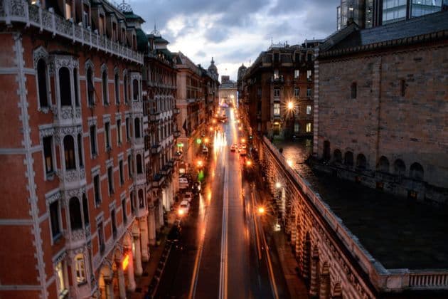 Una vista elevata di una strada cittadina bagnata al crepuscolo, con scie luminose di auto tra edifici storici sotto un cielo nuvoloso.