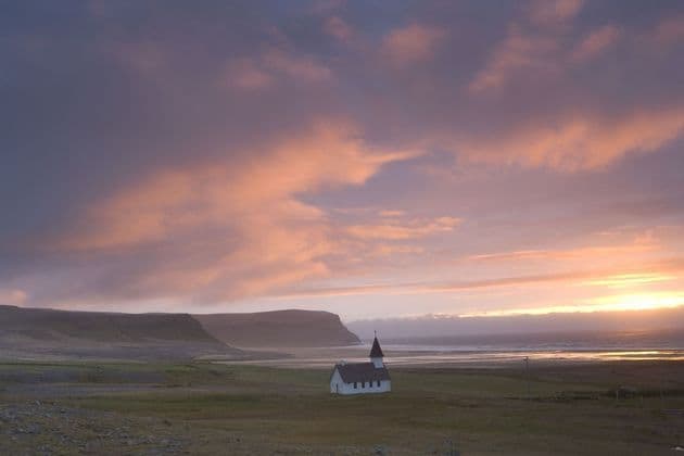 Eine kleine weiße Kirche steht einsam in einer grünen Landschaft vor einer Küste und Klippen unter einem farbenfrohen Sonnenuntergangshimmel.