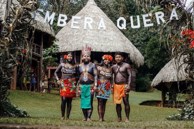 Vier Personen der Embera-Quera-Gemeinschaft in traditioneller Kleidung und Körperbemalung posieren vor Strohhütten.