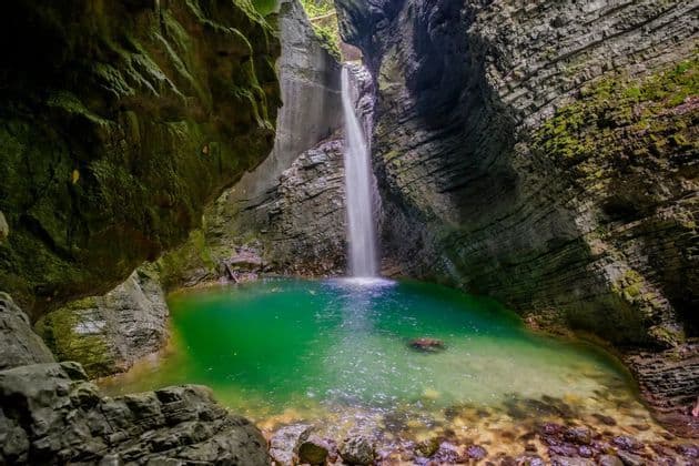 Ein Wasserfall stürzt in ein leuchtend türkisfarbenes Becken am Grund einer engen, moosbewachsenen Felsenschlucht.