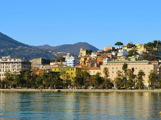Una vista sul lungomare di una città costiera con edifici colorati e palme, con verdi colline sullo sfondo sotto un cielo azzurro chiaro.