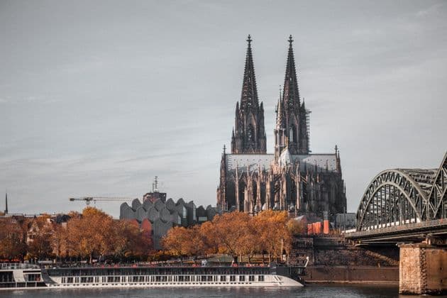 Un paesaggio urbano con una grande cattedrale gotica e un ponte ad arco visti da oltre un fiume, con alberi autunnali arancioni in primo piano.