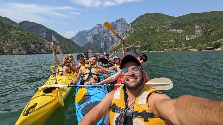 Un viaje en grupo de WeRoad se toma una selfie sonriente mientras hacen kayak en un lago con montañas verdes de fondo.