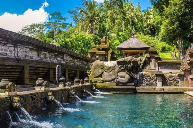 Verzierte Steinspeier speisen Wasser in ein türkisfarbenes Becken bei einem moosbewachsenen Tempel mit einer Elefantenskulptur, vor der Kulisse eines üppigen Dschungels.