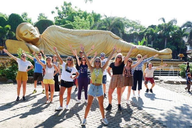 Un viaje en grupo de WeRoad posando con los brazos en alto frente a una estatua gigante y dorada de Buda reclinado.