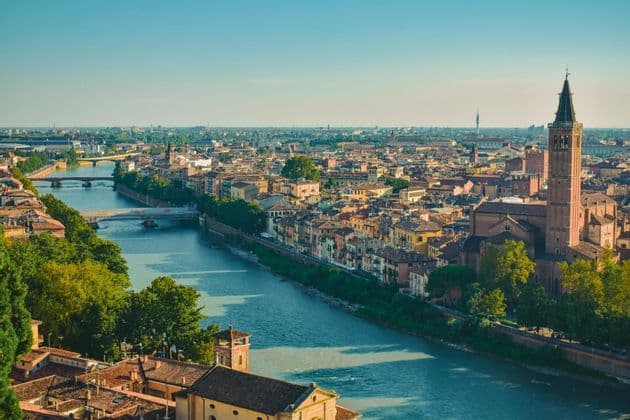 An aerial view of a historic city with a wide river, multiple bridges, terracotta roofs, and a prominent bell tower.