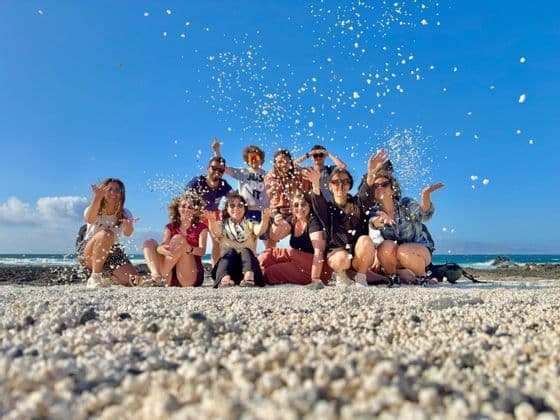 Un viaggio di gruppo WeRoad lancia sassolini bianchi in aria mentre posa per una foto su una spiaggia assolata.