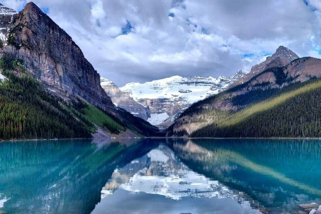 Snow-capped mountains and pine forests are reflected in the calm, turquoise water of an alpine lake under a cloudy sky.
