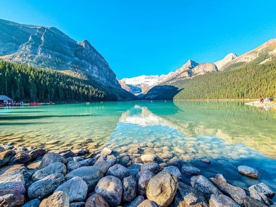 A clear turquoise lake reflects snow-capped mountains and pine forests under a blue sky, with a rocky shoreline in the foreground.